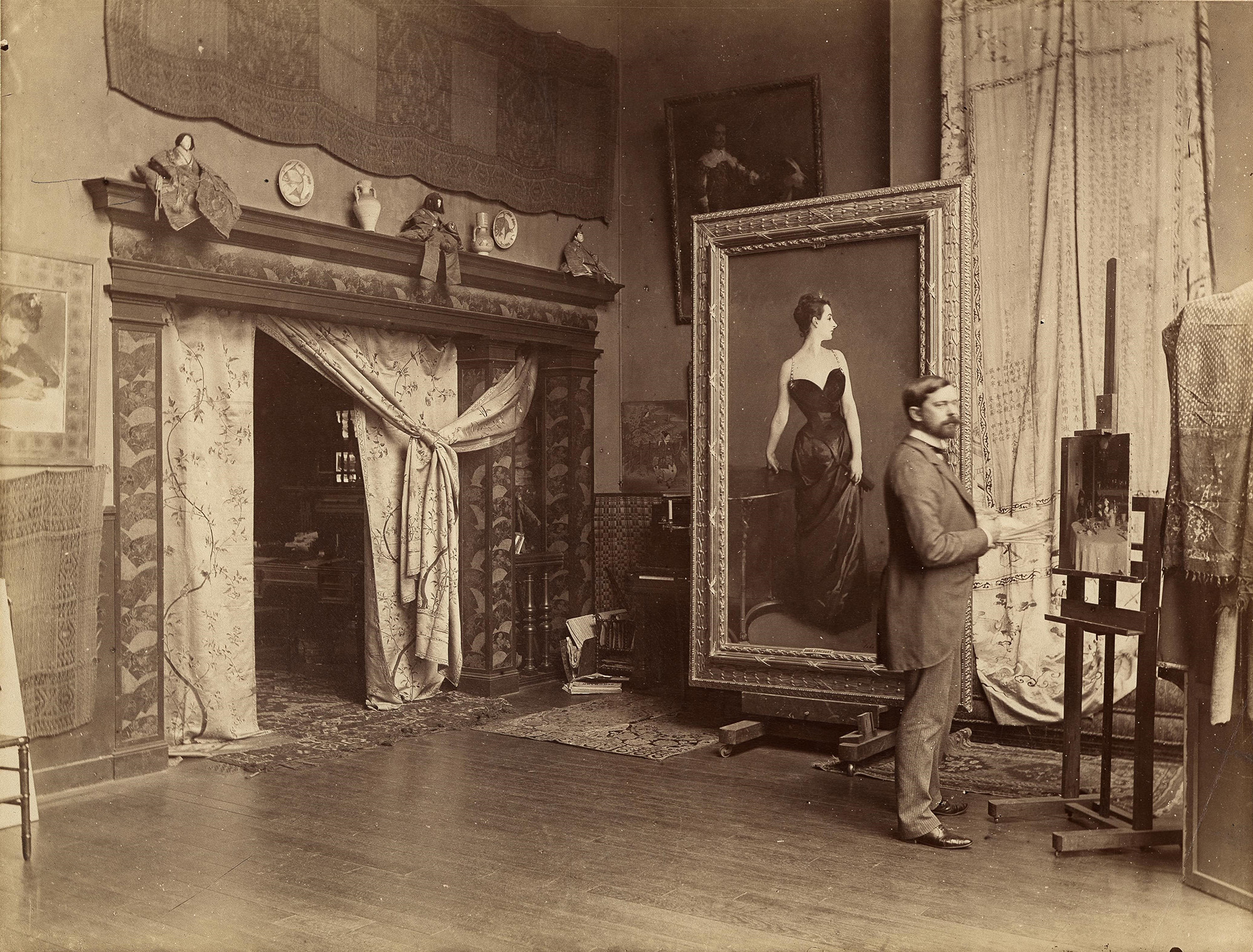 A historical photograph of master artist John Singer Sargent in his Paris studio, standing beside his iconic masterpiece portrait, "Madame X."
